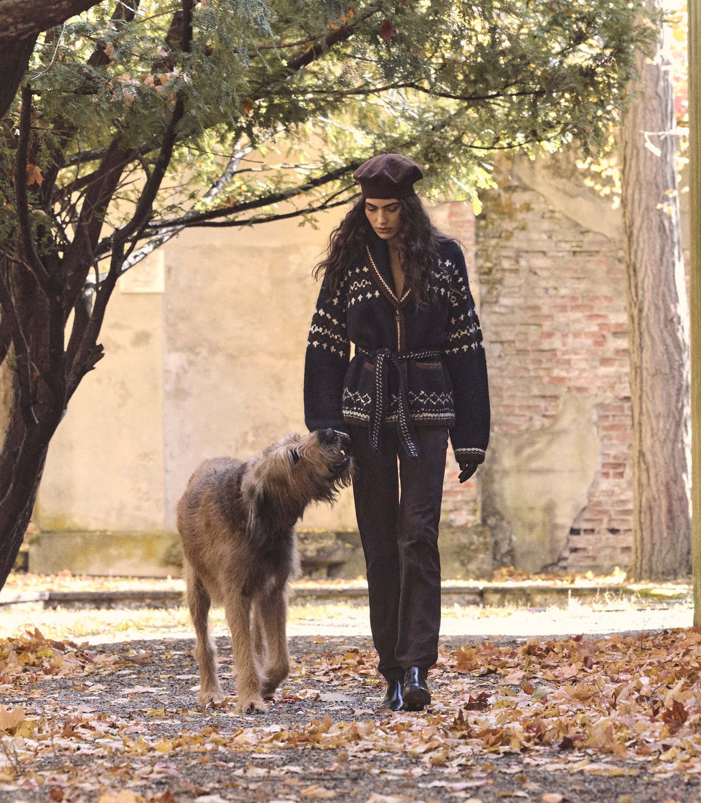 Woman in a patterned sweater and dark pants walking a dog in an outdoor setting with trees and a brick wall.