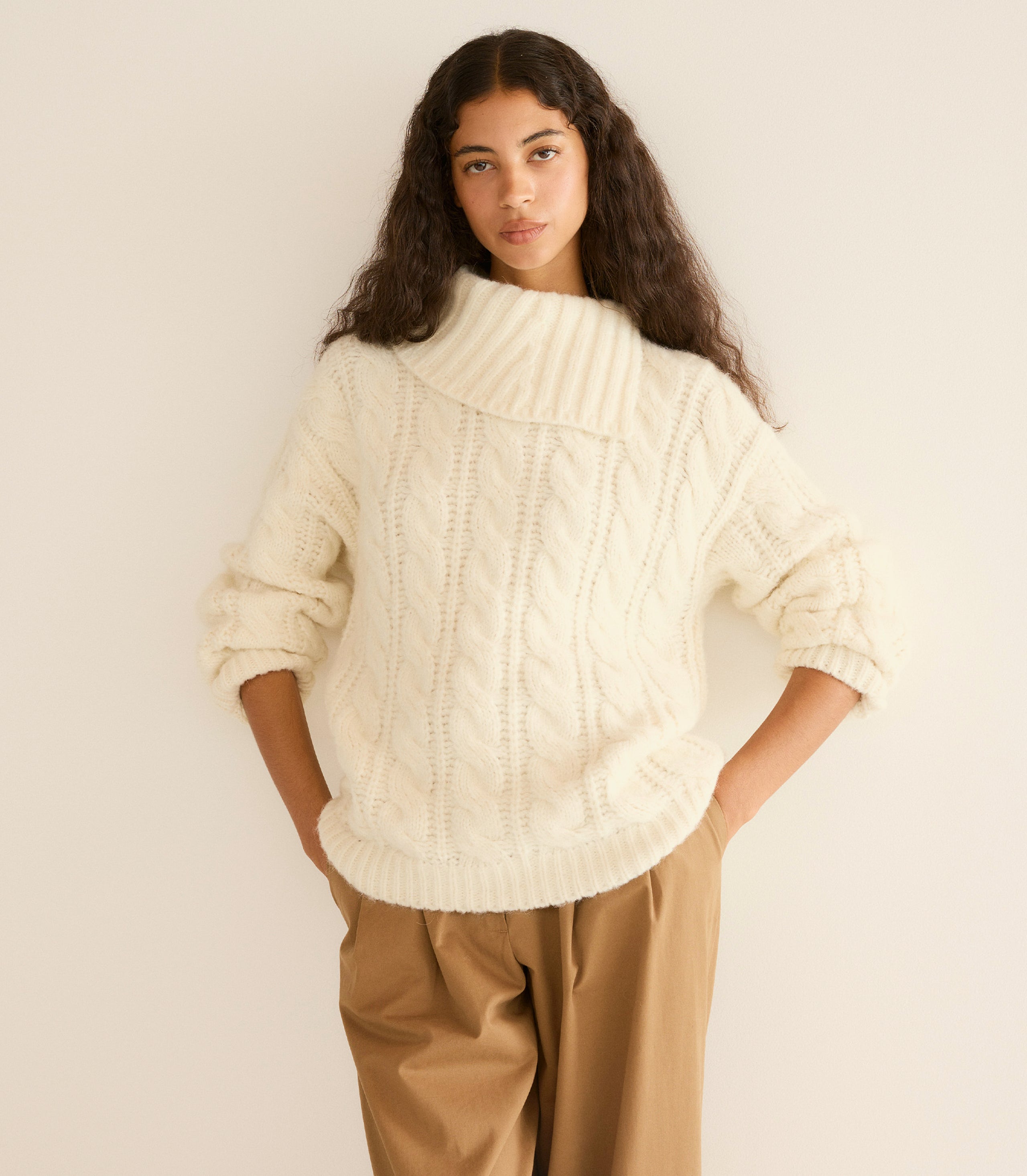 Woman wearing a cream sweater and beige pants against a plain background