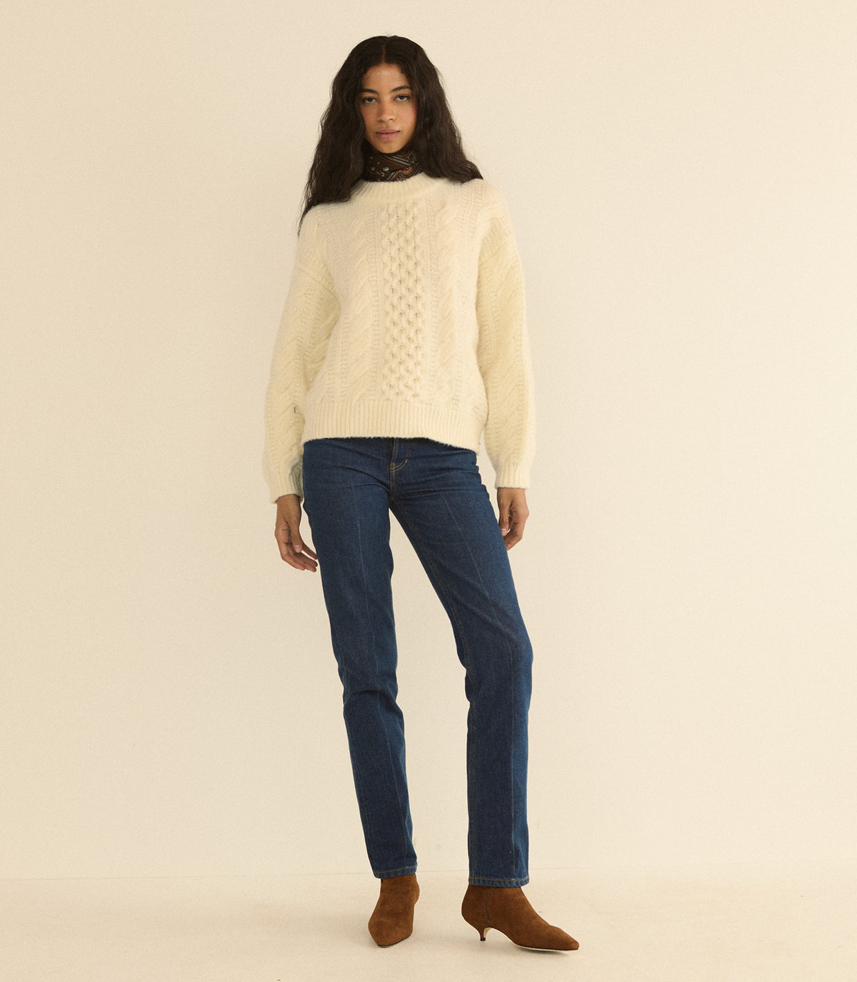 A woman modeling a cream cable-knit mock-neck sweater, paired with dark blue jeans and brown ankle boots, against a solid warm beige studio background.