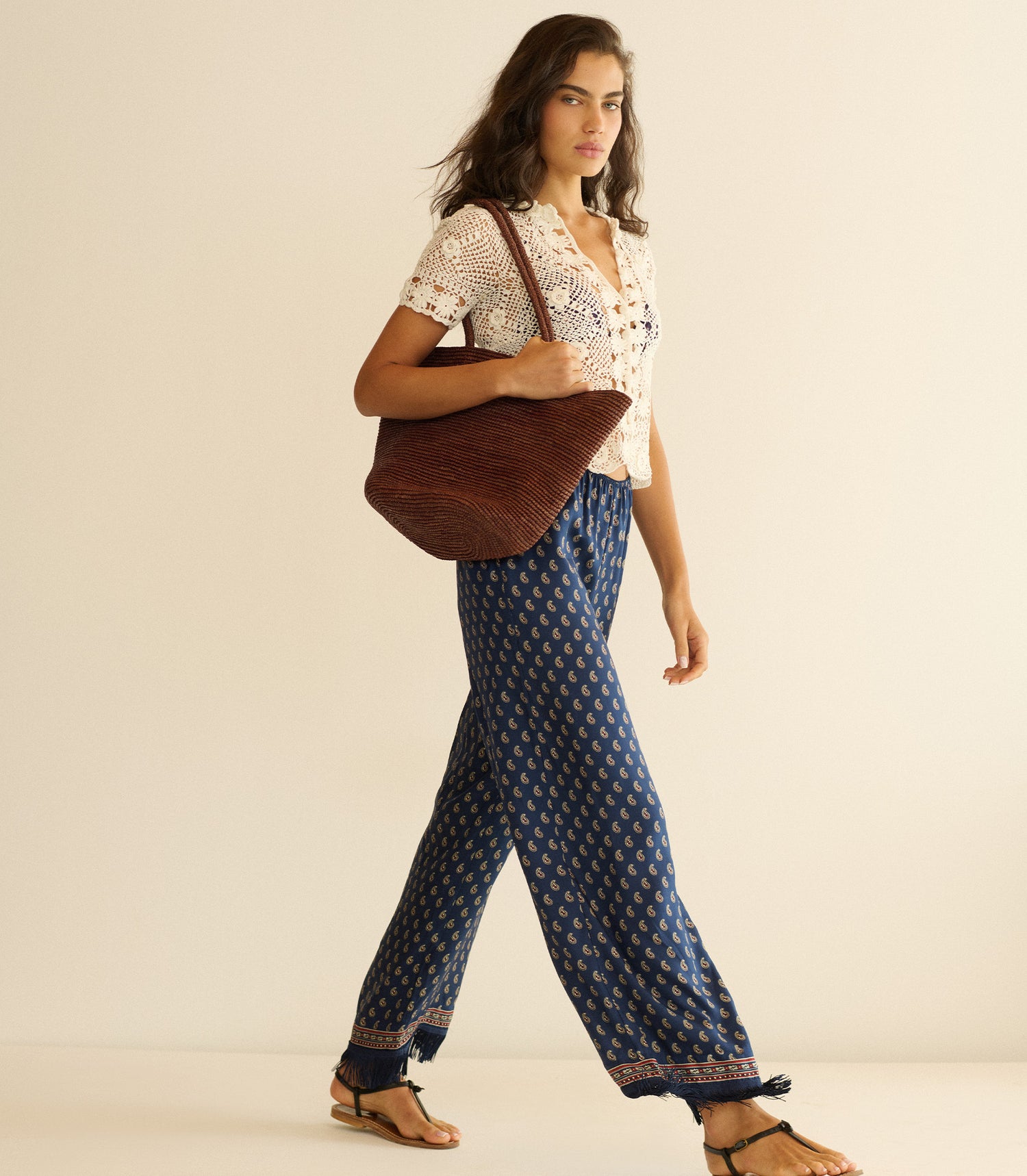 Woman wearing a white blouse and blue patterned pants, holding a brown tote bag on a beige background