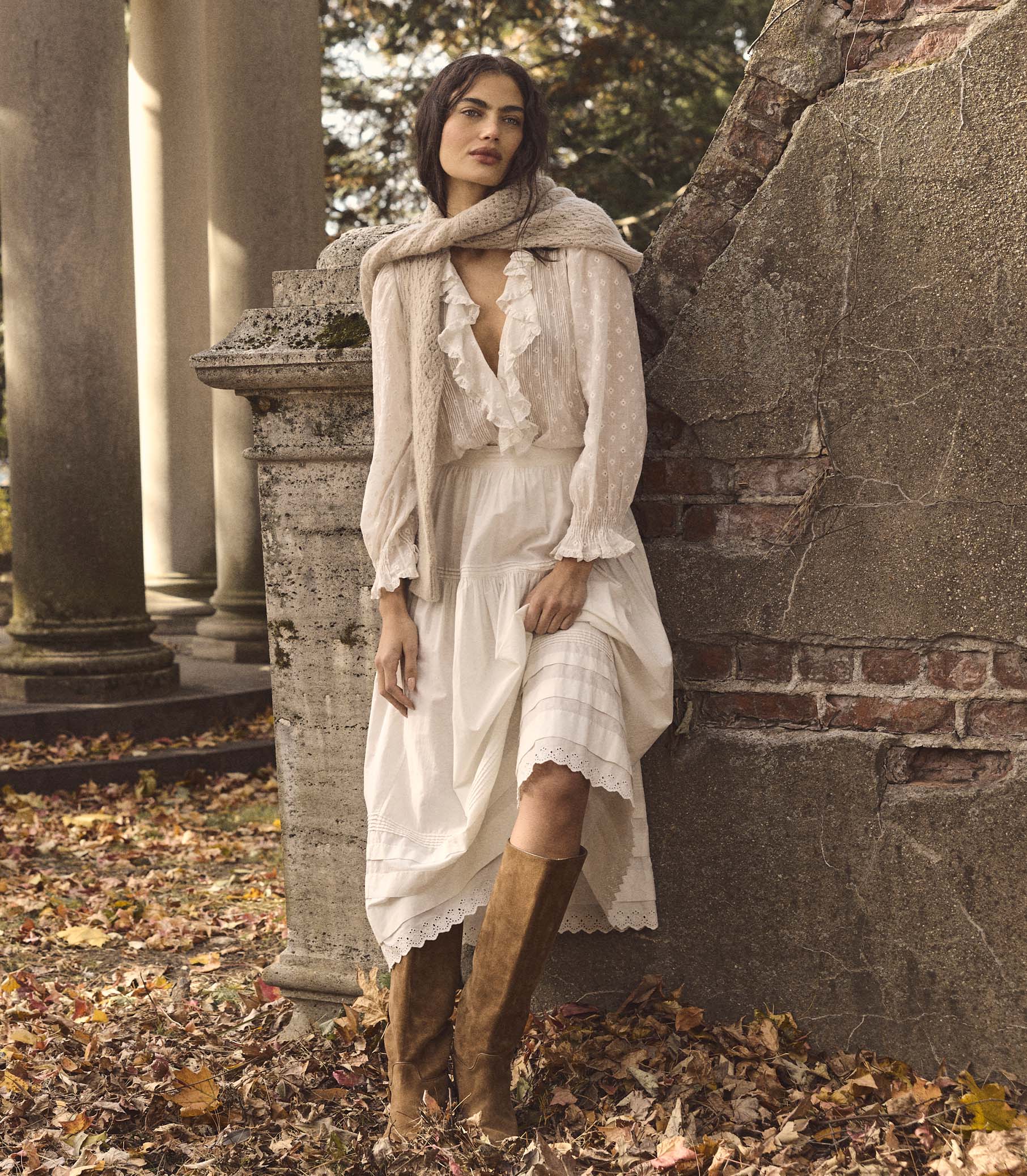 Woman in a white blouse, white skirt, and brown boots standing in an autumnal setting with stone architecture
