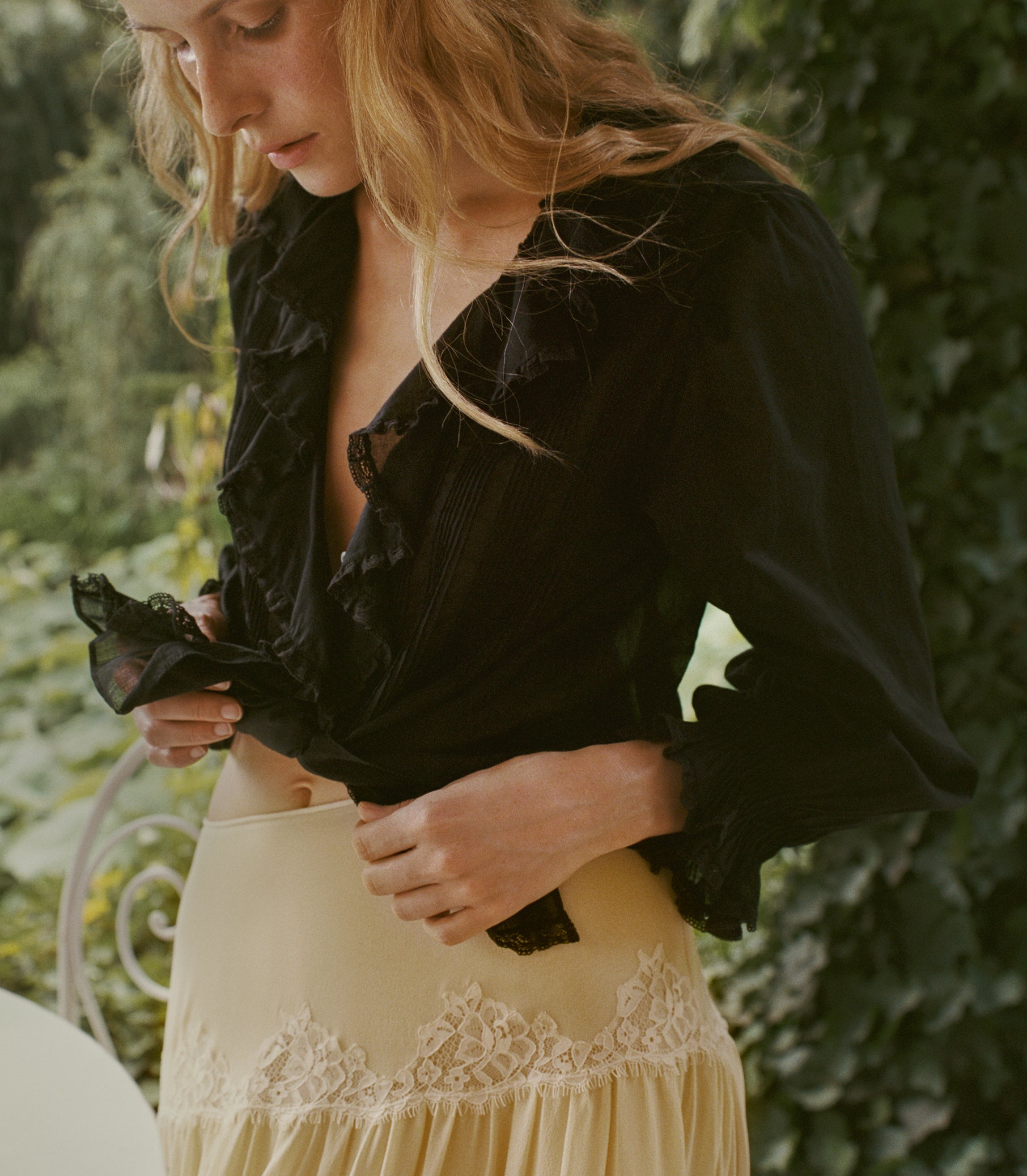 A woman outdoors wearing a black ruffled blouse and a cream lace-trimmed skirt, with green foliage in the background.