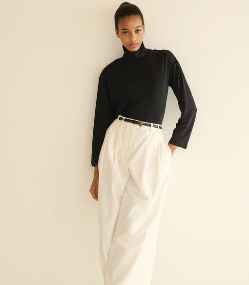 Person wearing a black turtleneck and white pants against a plain background