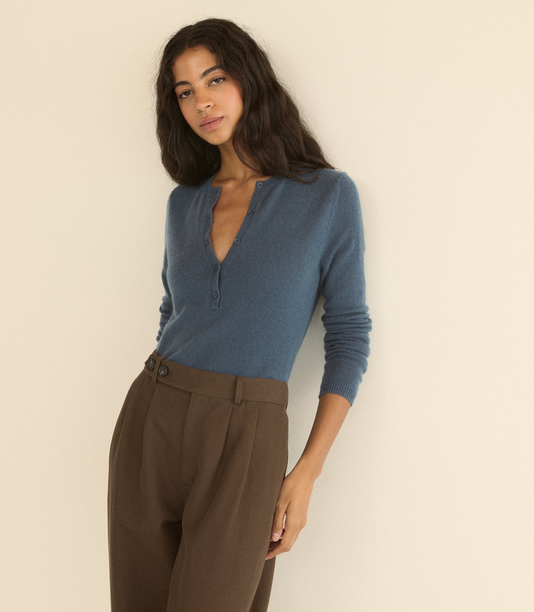 A woman wearing a dusty blue long-sleeve henley top and brown trousers, posing against a plain beige wall. 1
