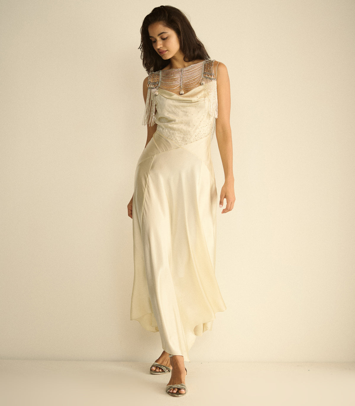 A woman modeling a cream-colored satin maxi dress with a beaded lace bodice and sheer straps, standing against a plain cream studio background. 1
