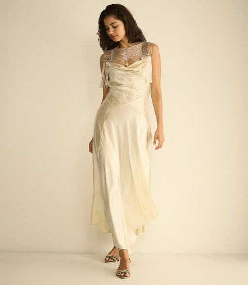 A woman modeling a cream-colored satin maxi dress with a beaded lace bodice and sheer straps, standing against a plain cream studio background.