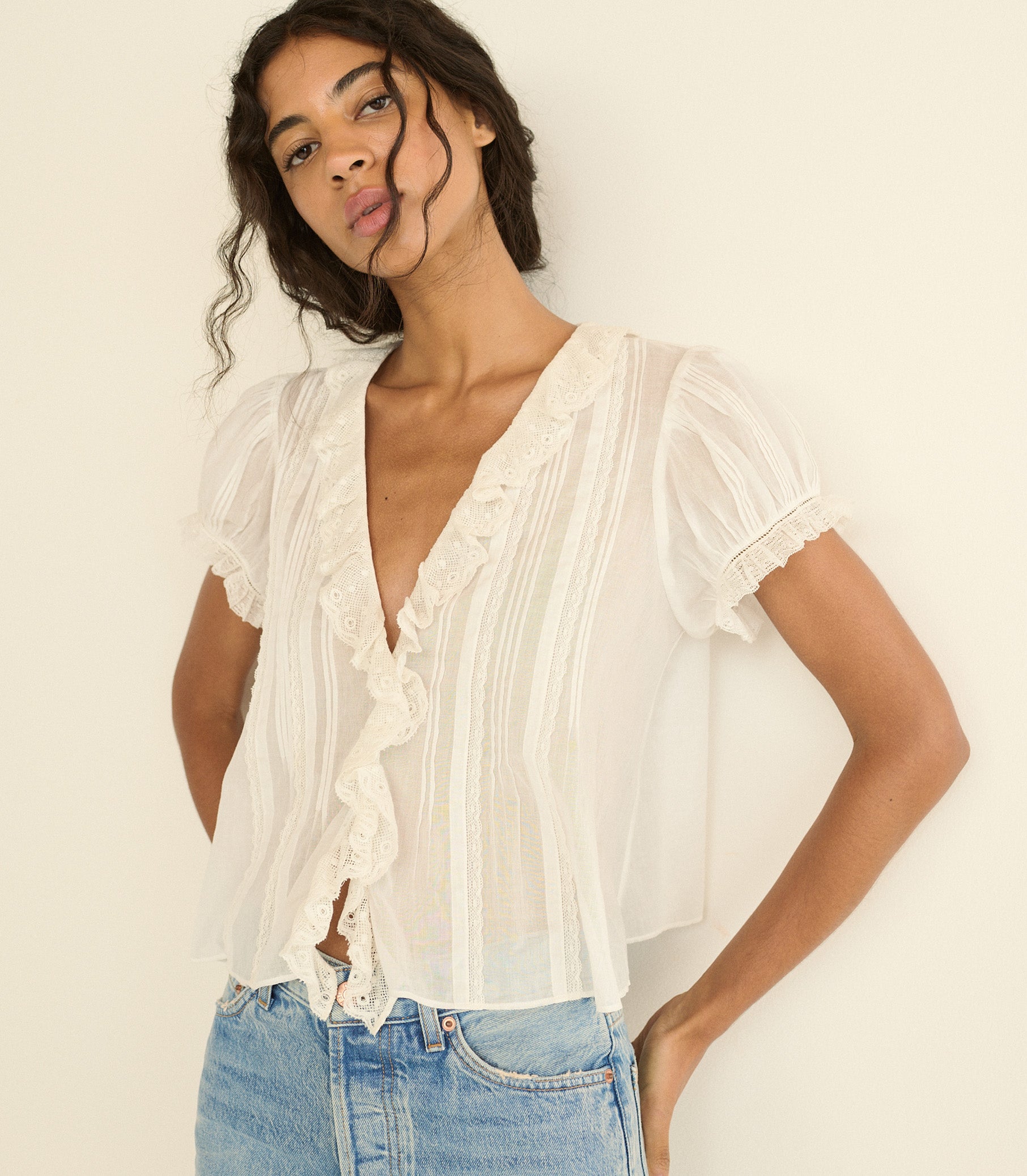 A woman wearing a sheer white short-sleeve blouse with ruffle trim and vertical pleats, paired with light blue jeans, posing against a plain off-white background.