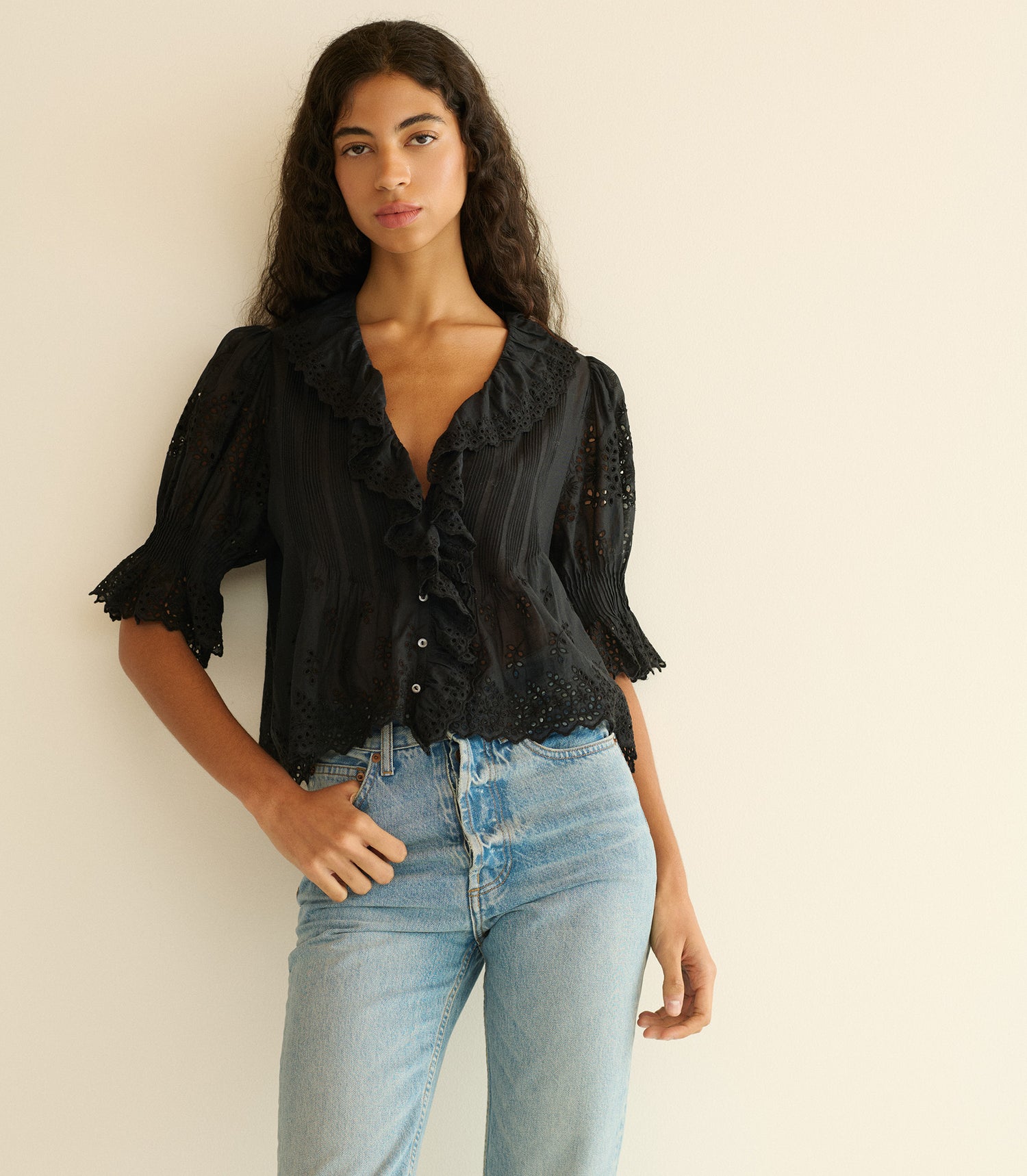 Woman wearing a black blouse with ruffled details and light blue jeans against a beige wall.