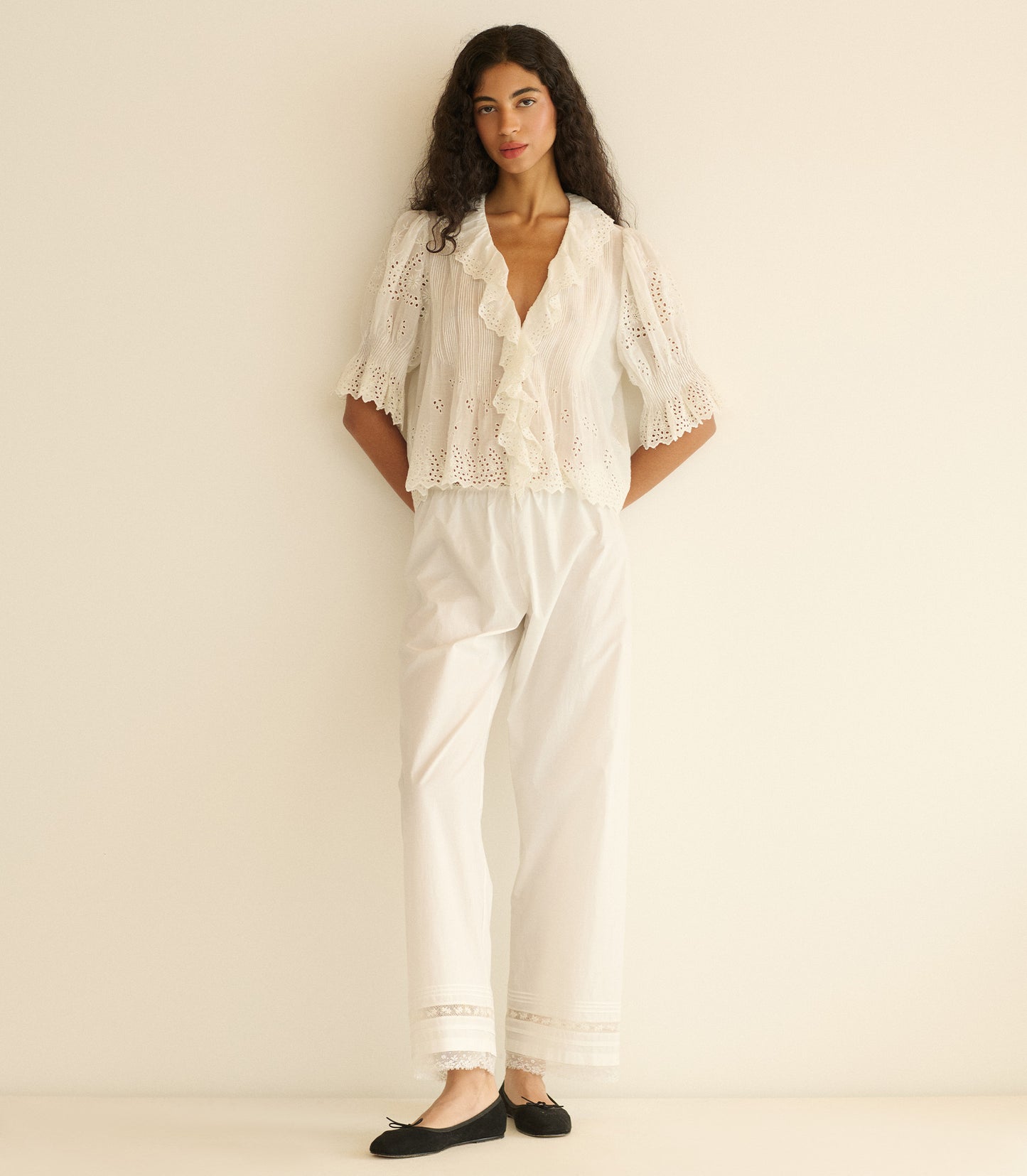 Woman wearing a white eyelet blouse and white pants with lace details on a plain background