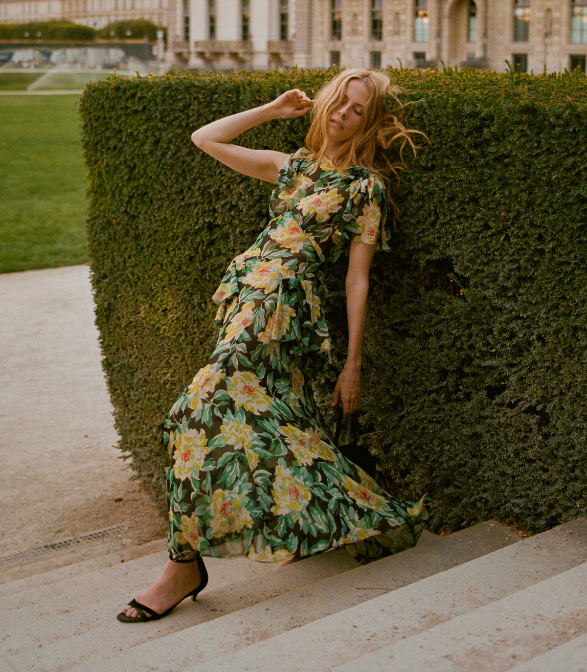 A woman posing on stone steps in front of a large green hedge, wearing a dark green tiered maxi dress with a yellow and orange floral print and black pointed-toe flats.