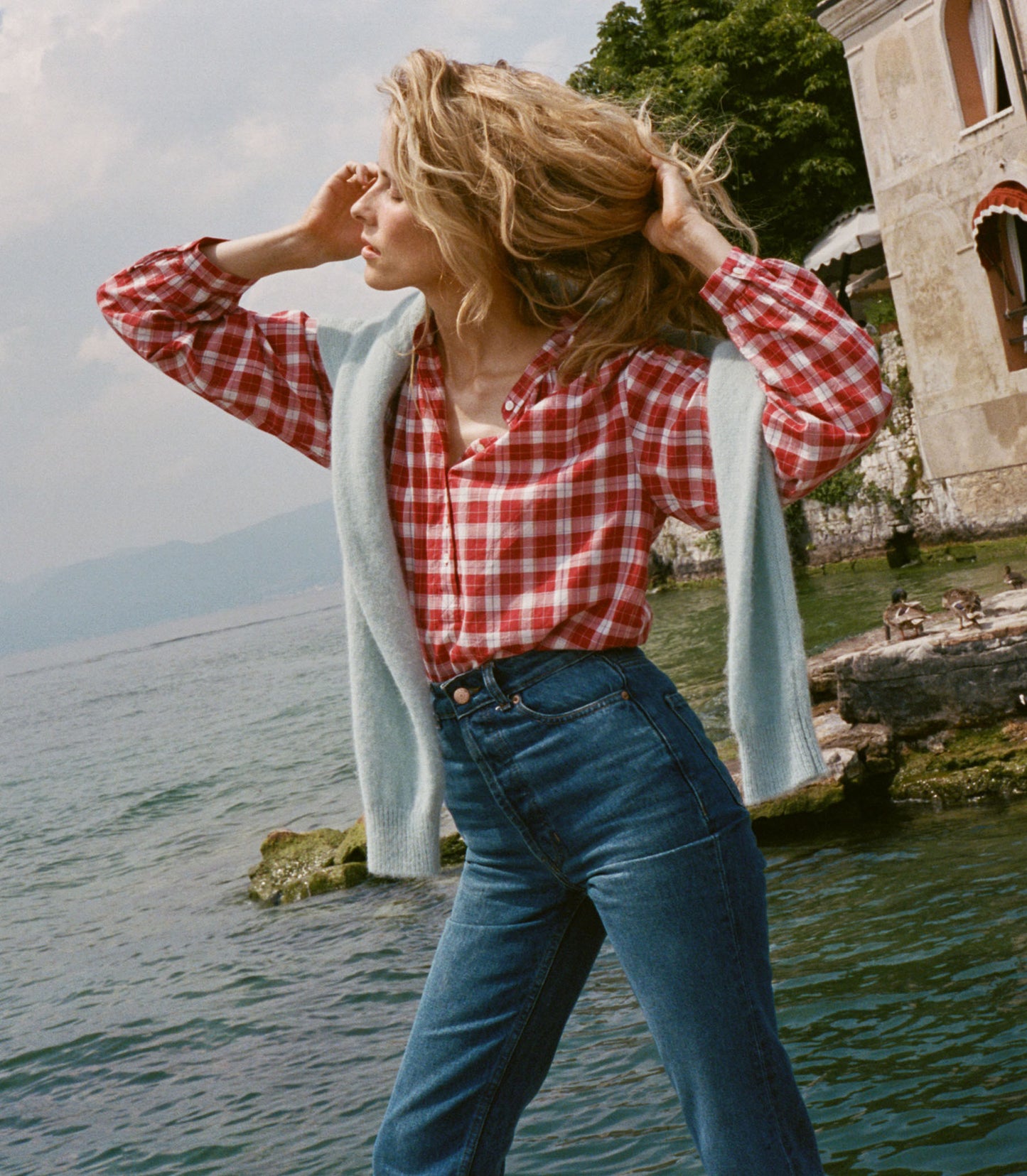 Woman in a red checkered shirt and blue jeans standing by a body of water with a scenic background.