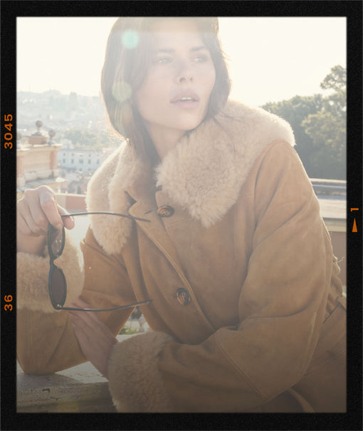 A woman wearing a tan shearling-lined coat with a large cream fur collar, holding black sunglasses, set against a warm, sunlit outdoor background with a black film border.
