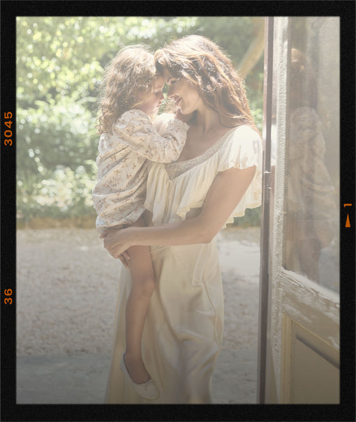 A woman in a cream-colored dress holds a young child in a matching cream floral top, standing by an open door in a sunlit outdoor setting with a black film-style border overlay.