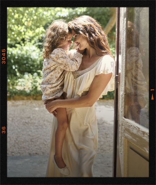 A woman in a cream satin slip dress holds a young child wearing a matching cream floral long-sleeve top, standing in a doorway with a warm, sunny outdoor background.