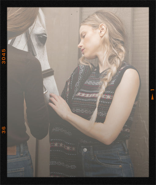 A woman in a sleeveless patterned knit top and blue jeans stands beside a horse, touching its neck, with another person partially visible on the left. The photo has a vintage film-strip border effect.
