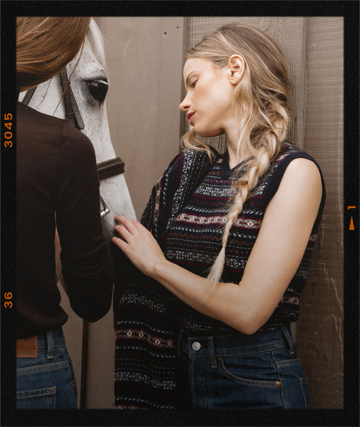 A woman in a sleeveless patterned knit top and blue jeans stands beside a horse, touching its neck, with another person partially visible on the left. The photo has a vintage film-strip border effect.