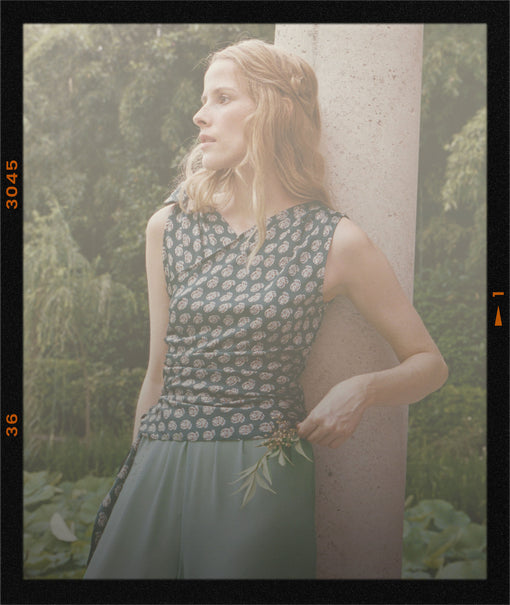 A woman wearing a sleeveless patterned top and muted green bottoms leans against a stone pillar in a garden setting, framed by a black border with orange film-date markings.