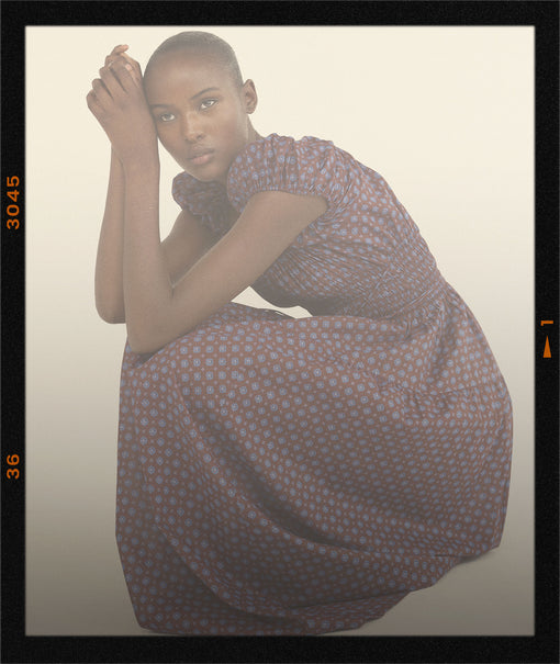 A model in a short-sleeved, knee-length dress with a small-scale geometric pattern in muted purple and orange, posed in a crouching position against a light beige background with a black border.