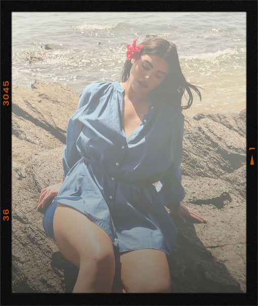 Woman in a blue dress sitting on a rocky beach with a red flower in her hair.