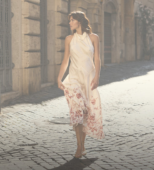 A woman walks down a sunlit cobblestone street wearing a sleeveless cream halter dress with a red floral print concentrated along the hem.
