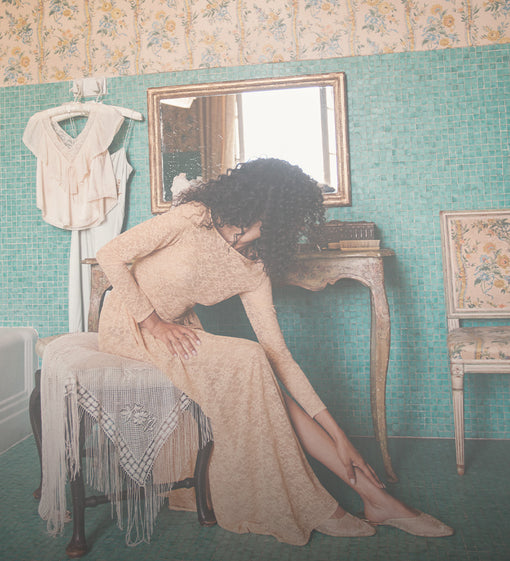 A woman with curly dark hair sits on a chair in a vintage-style room with turquoise mosaic tile walls, wearing a beige lace long-sleeve maxi dress and nude heels. A white blouse hangs on a hanger on the left, and a mirror, console table, and floral chair are visible.