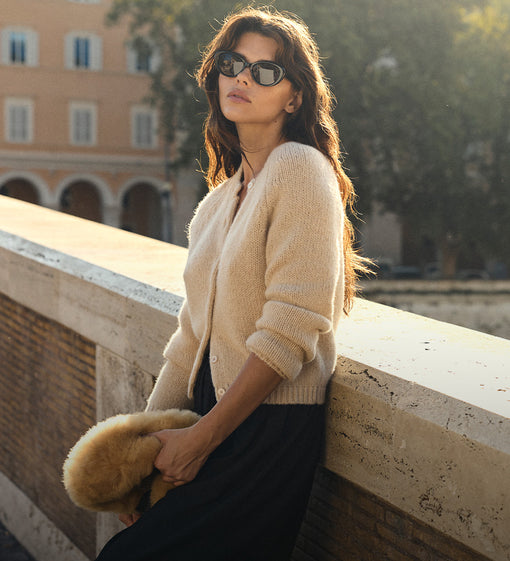 A woman wearing a cream-colored knit cardigan, black skirt, and sunglasses leans against a stone wall in an outdoor setting with warm sunlight.