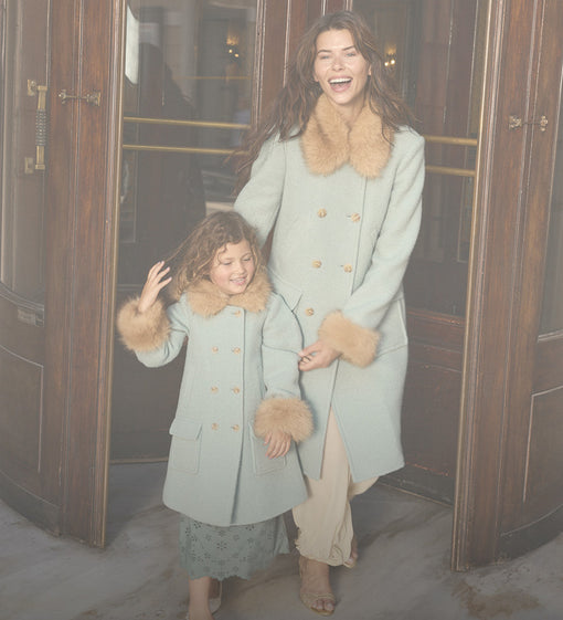A woman and a young girl wearing matching sage green wool coats with tan faux fur trim and gold buttons, standing in front of dark wooden doors.