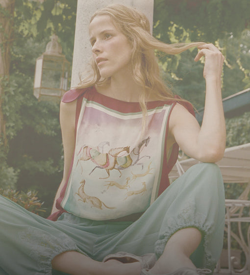 A woman sitting outdoors wearing a sleeveless top with a horse and rider illustration and light green pants, photographed in a softly filtered, vintage-style outdoor scene.