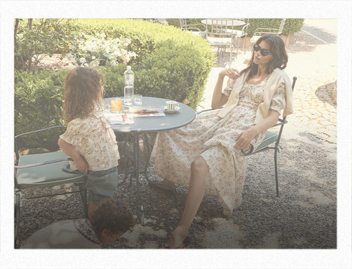 A woman and a child wearing matching cream floral print dresses sit at an outdoor patio table, with another person visible in the foreground. The scene is shot in a warm, sunlit, vintage-filtered style with a white border.