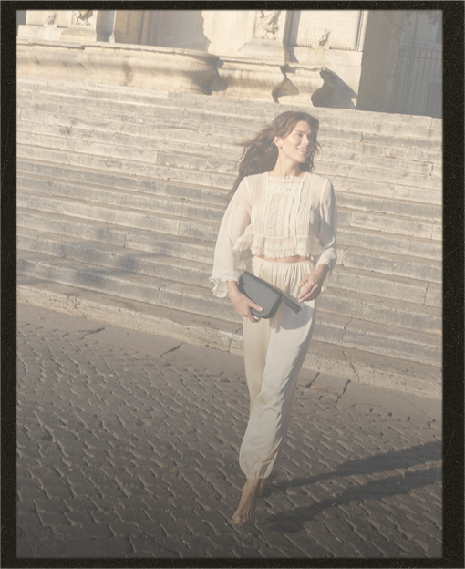 A woman walking outdoors on stone steps and a cobblestone street, wearing a light cream or off-white long-sleeve top and matching pants, holding a dark clutch bag.