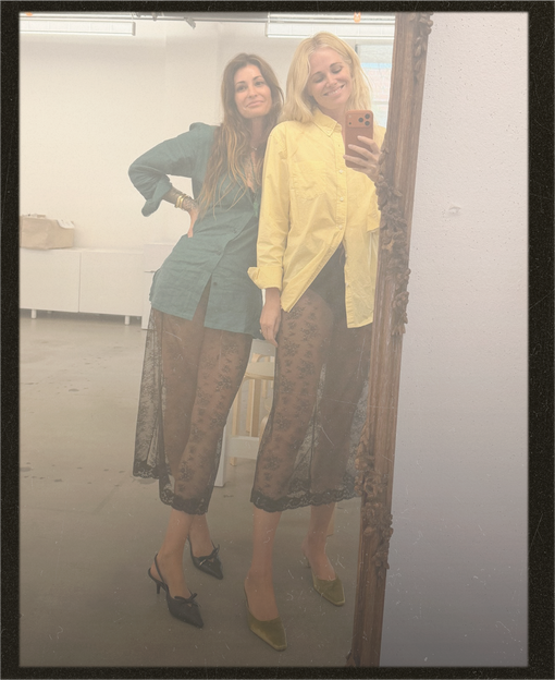 Two women taking a mirror selfie in a bright studio space. The woman on the left wears a teal long-sleeve top and sheer brown patterned cropped pants with black heels.
