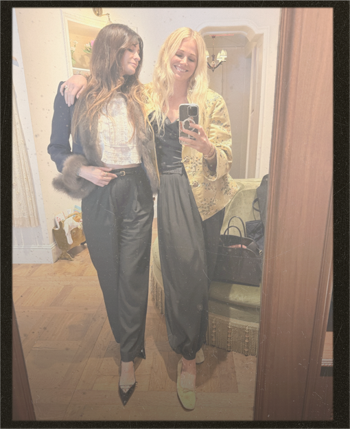 Two women taking a mirror selfie in a room with wood floors. The woman on the left wears a dark jacket with a fur collar over a white top and dark pants.