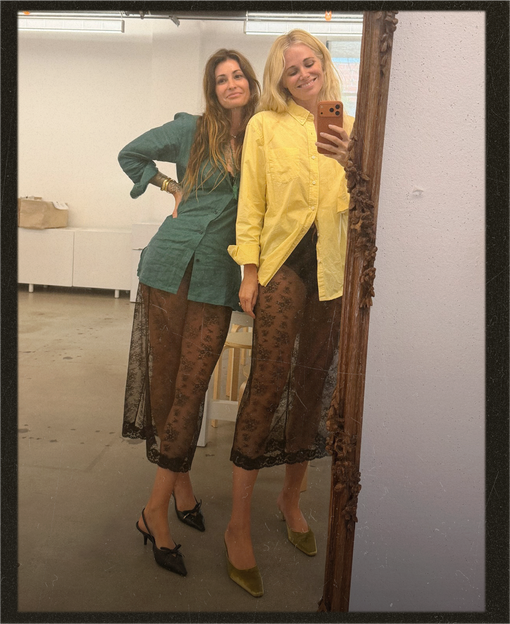 Two women taking a mirror selfie in a studio setting, showcasing different colorways of a matching two-piece outfit. One outfit is teal green with brown patterned pants, and the other is yellow with brown patterned pants.