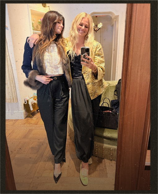 Two women taking a mirror selfie in a cozy indoor setting. The woman on the left wears a dark navy cardigan with a brown fur-trim, a white lace top, and black satin trousers.