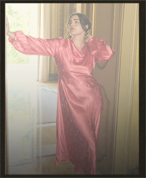 A woman wearing a shiny pink satin maxi dress with long balloon sleeves, posing in an indoor setting with a window and gold-toned door frame.