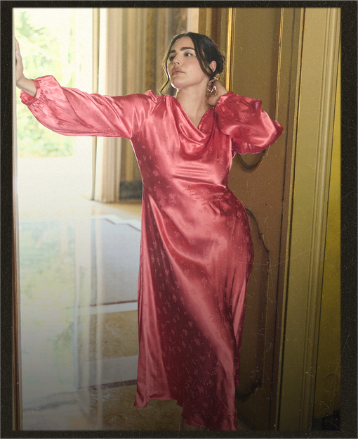 A woman wearing a shiny pink satin maxi dress with long balloon sleeves, posing in an indoor setting with a window and gold-toned door frame.