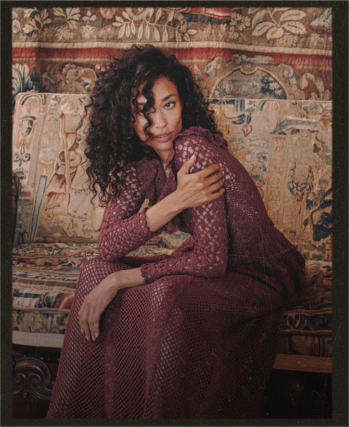 A woman with long curly dark hair sits on an ornate tapestry-covered sofa, wearing a long-sleeve burgundy textured knit dress or robe. The background is a detailed multicolored tapestry with figures and floral motifs.