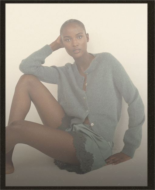 A model wearing agreen button-up cardigan and matching green shorts with dark lace trim, posed against a warm off-white background.