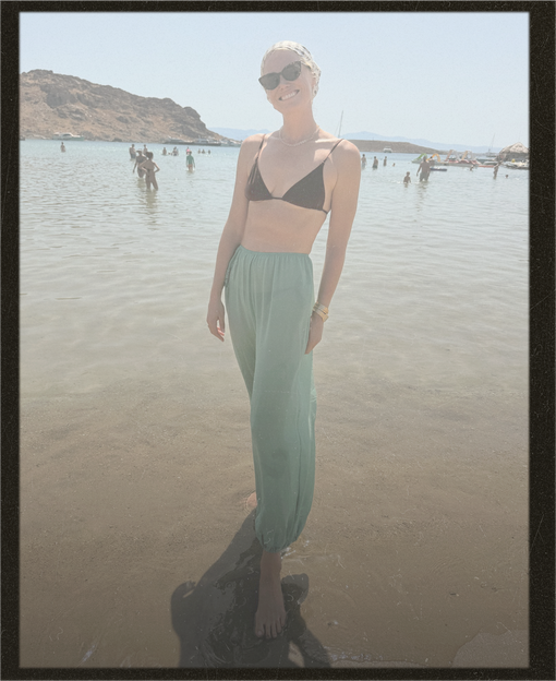 A woman stands in shallow water at a beach wearing a black triangle bikini top and light sage green high-waisted cropped pants. She is also wearing dark sunglasses and bracelets.
