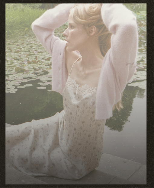 A woman in a pale pink cardigan and a white floral slip dress sits by a pond with lily pads, looking off-camera. The image has a soft, vintage, slightly faded aesthetic with a black border.
