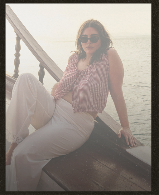 Woman sitting on a boat wearing sunglasses and a red patterned top with white pants.