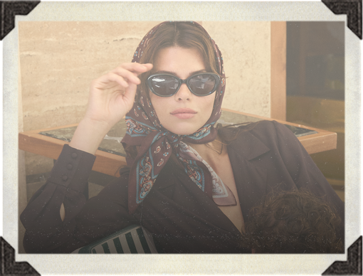A woman wearing dark sunglasses with a paisley-patterned headscarf and a dark blazer, posed in front of a rustic wooden table.