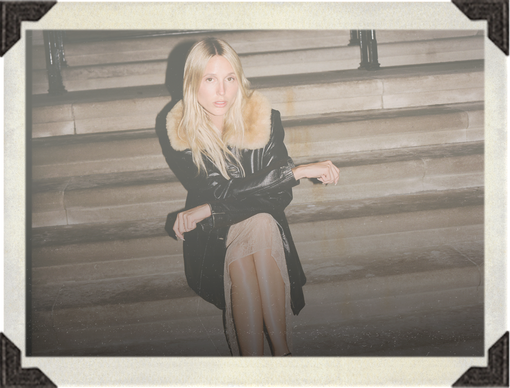A blonde woman sits on stone steps wearing a black coat with a beige fur collar, a beige skirt, and sheer black tights. The photo has a vintage Polaroid-style border with a warm cream background.
