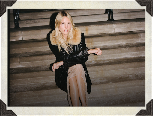 A woman with blonde hair sits on stone steps wearing a black coat with a large tan fur collar, sheer nude tights, and a black top. The photo has a vintage Polaroid-style border.