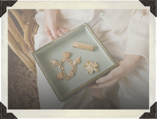 A person holds a square display tray containing three small vintage-style gold-toned jewelry pieces, including a floral brooch, a chain motif piece, and a small rectangular charm, set against a soft cream-colored outfit and warm neutral background.