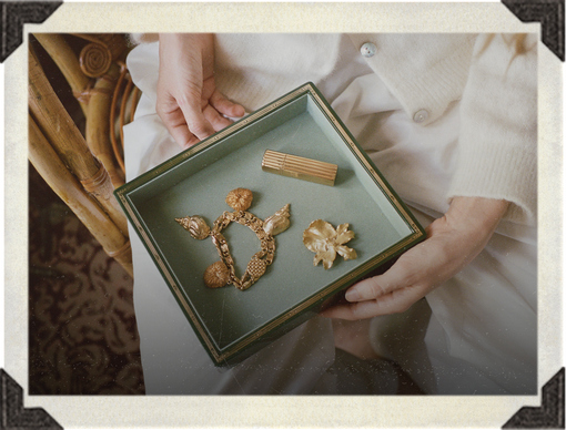 A person in a cream-colored cardigan holds a square green jewelry box containing three vintage gold-toned brooches, photographed with a decorative border overlay.