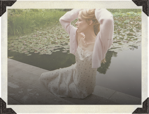 A woman in a white lace slip dress and pale pink cardigan sits by a pond with lily pads, framed by a vintage-style cream border with black corner details.