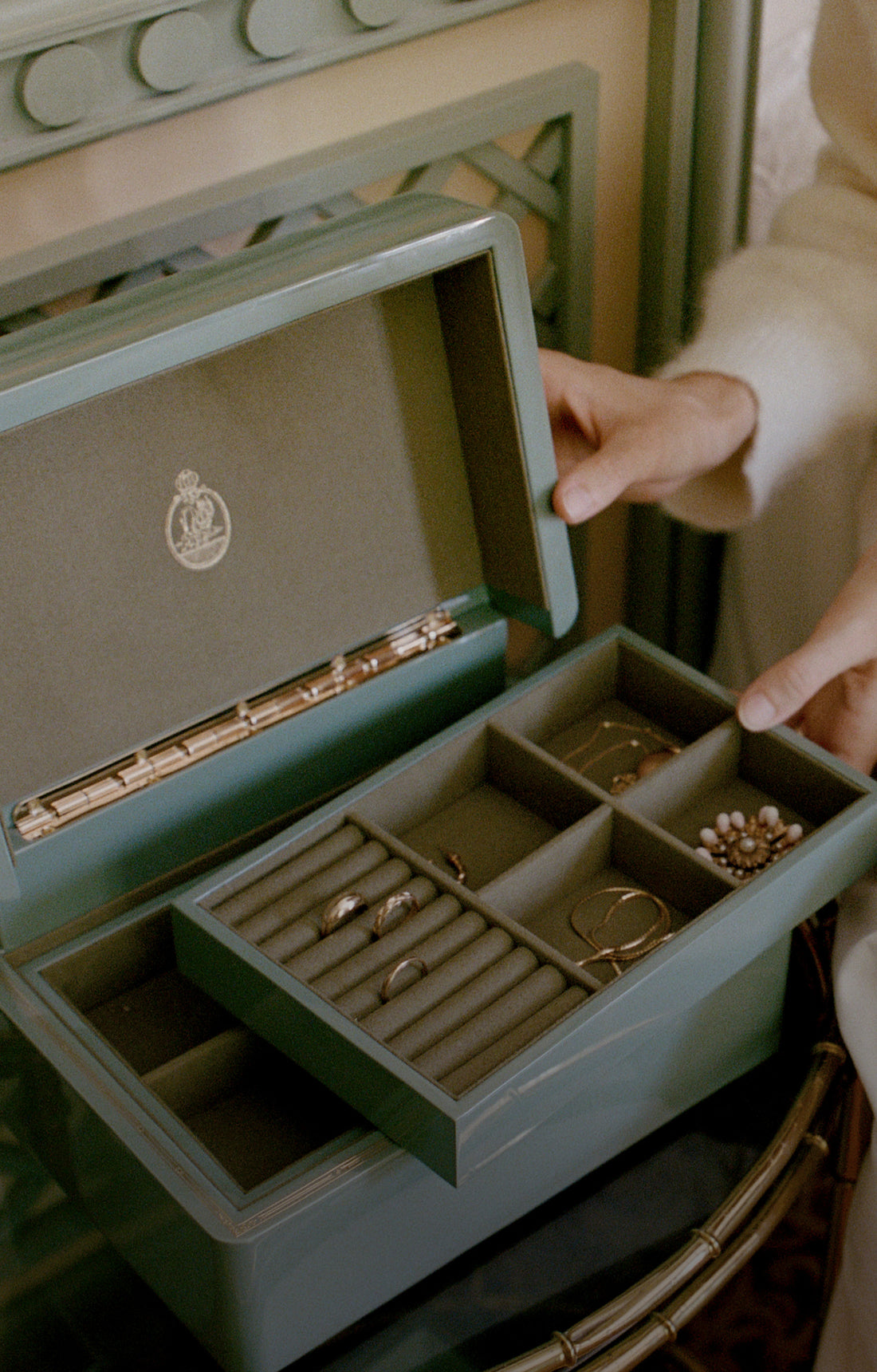 A sage green jewelry box with a sliding drawer pulled open, displaying various gold and pearl jewelry pieces inside. A person's hands are visible holding the box and pulling the drawer, set against a softly blurred background.