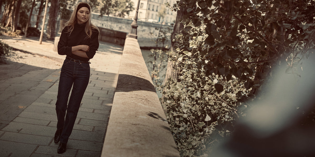 A woman wearing a black long-sleeve top and dark jeans walks along a stone pathway beside a canal with dense foliage on the right.