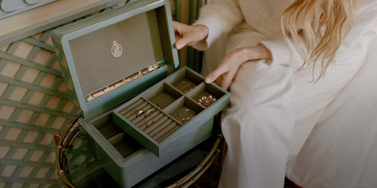 A woman in a white robe opens a sage green jewelry box containing rings and necklaces, set on a woven table.